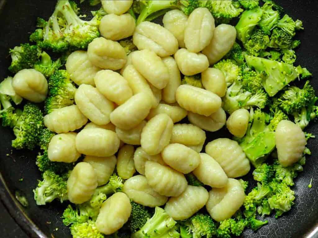 Close-up of uncooked gnocchi pasta piled on top of chopped broccoli in a black frying pan. The broccoli is bright green and the gnocchi is a light beige color, showcasing a fresh and colorful combination.